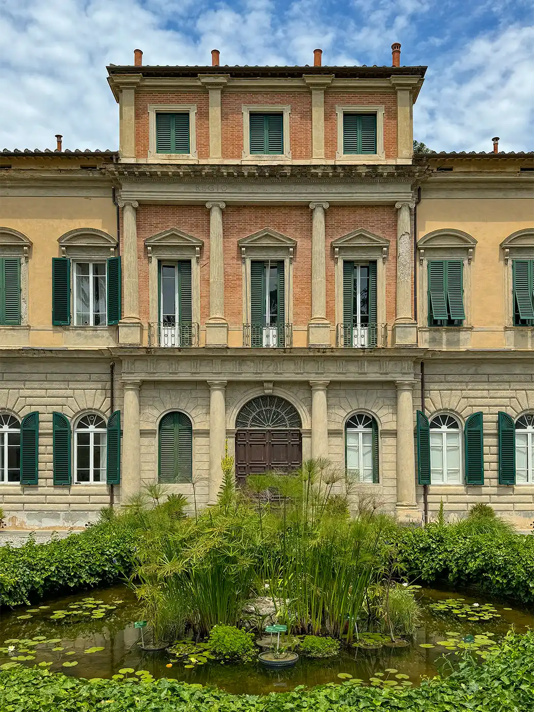 Geheimtipp-Pisa-Botanischer-Garten Eine alte Villa mit grünen Fensterläden, Säulen und einem kleinen Teich mit Pflanzen davor, unter einem teilweise bewölkten Himmel.