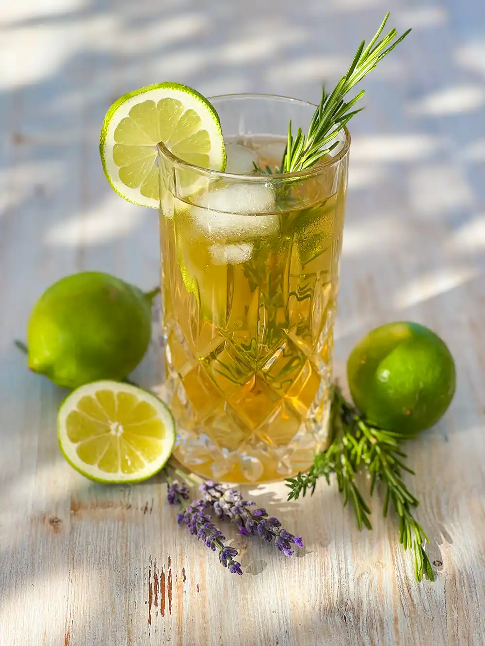 Ein gekühltes Kräutergetränk mit Limettenscheiben, Rosmarin und Lavendel auf einem Holztisch, daneben liegen ganze Limetten neben dem Glas.