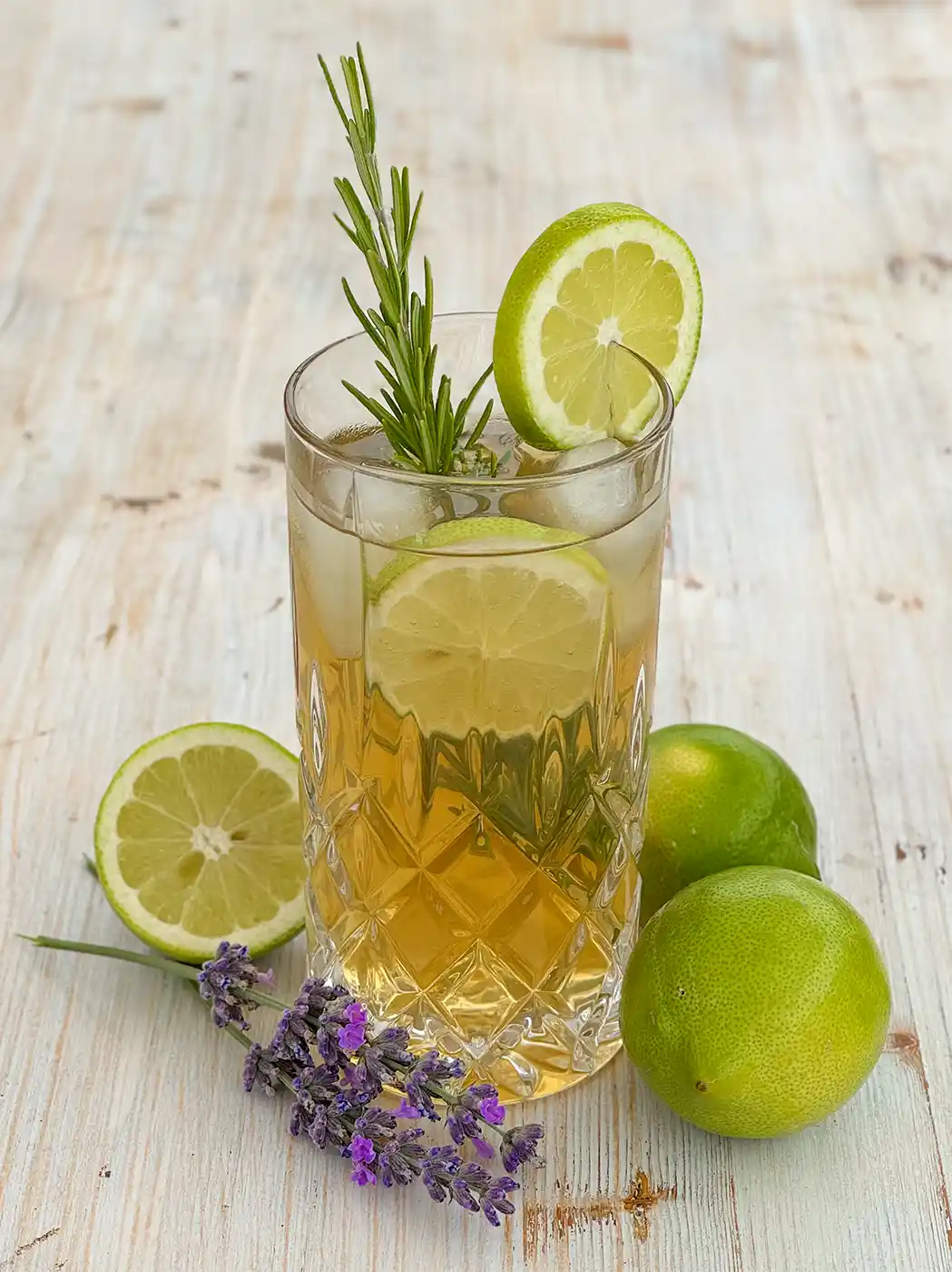 Ein gekühltes Kräutergetränk mit Limettenscheiben, Rosmarin und Lavendel auf einem Holztisch, daneben liegen ganze Limetten neben dem Glas.