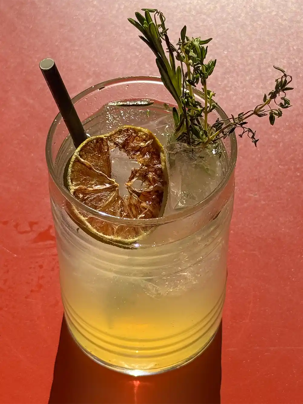 Ein Cocktail mit Eis, einem Pappstrohhalm, garniert mit getrockneter Limette und Kräutern, auf rotem Hintergrund.