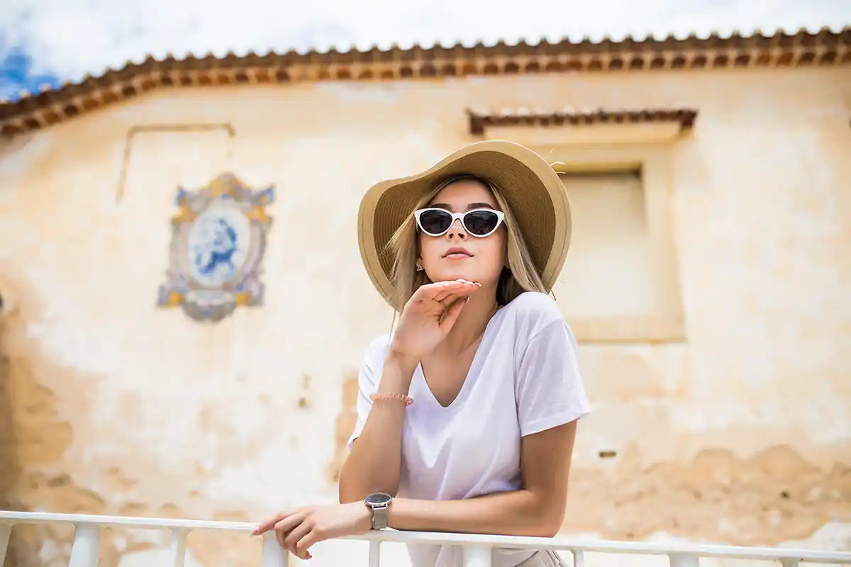 Eine Frau mit Sonnenbrille und Sonnenhut lehnt an einem weißen Geländer, im Hintergrund ist ein altes, beiges Gebäude zu sehen.