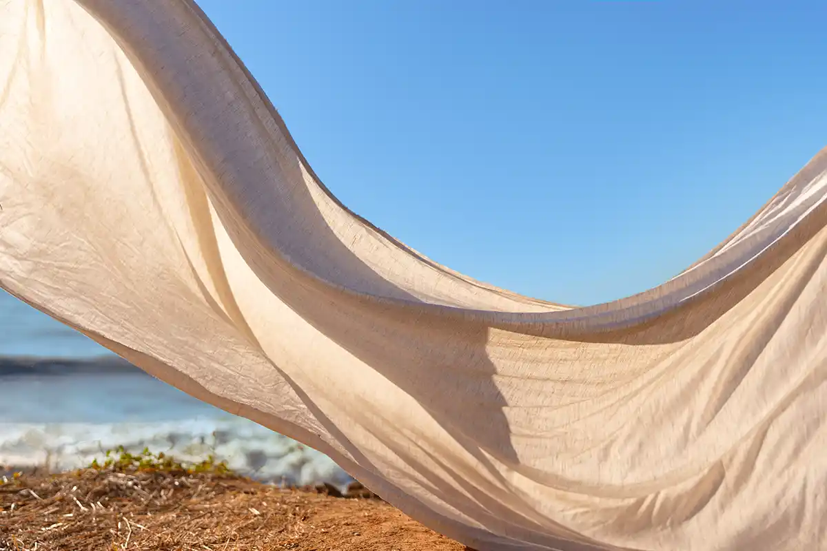 Beiger Stoff, der draußen im Wind weht, im Hintergrund blauer Himmel und Meereswellen.
