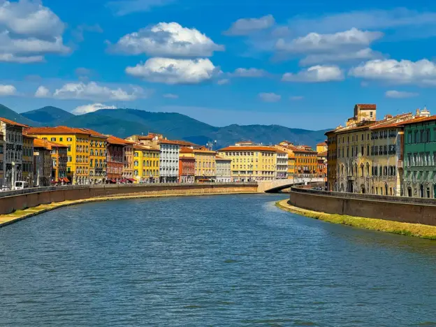Bunte Gebäude säumen einen Fluss unter blauem Himmel, im Hintergrund sind Wolken und ferne Berge zu sehen.