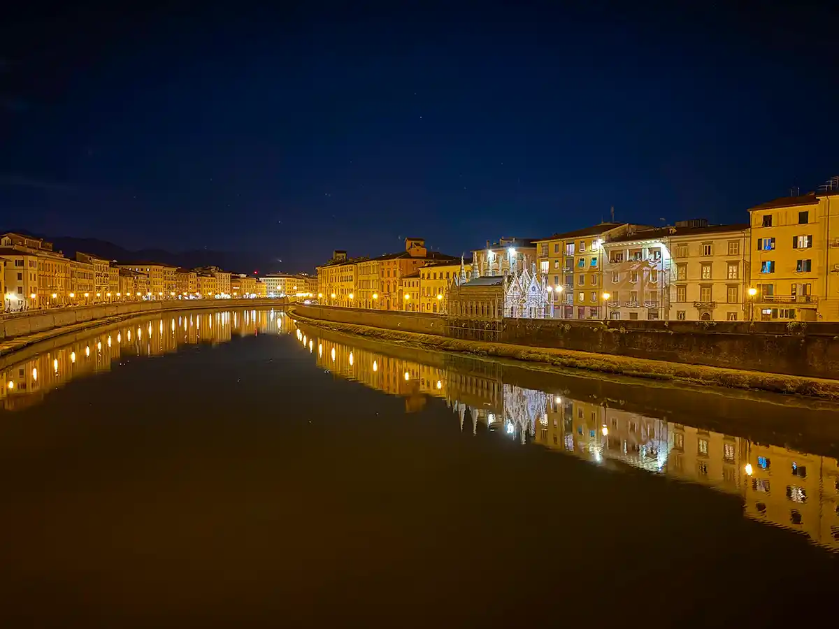 Pisa-Arno-Ufer-Abend Ein ruhiger Fluss spiegelt nachts unter dem dunkelblauen Himmel die beleuchteten historischen Gebäude und eine kleine Kirche wider.