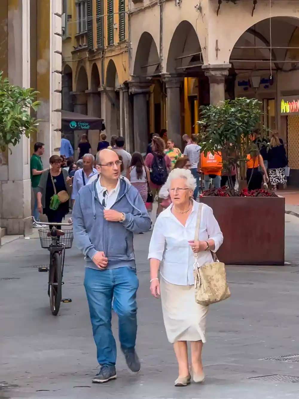 Im Hintergrund sind eine ältere Frau und ein Mann zu sehen, die eine belebte Straße entlanggehen, die von Geschäften und Menschen gesäumt ist.