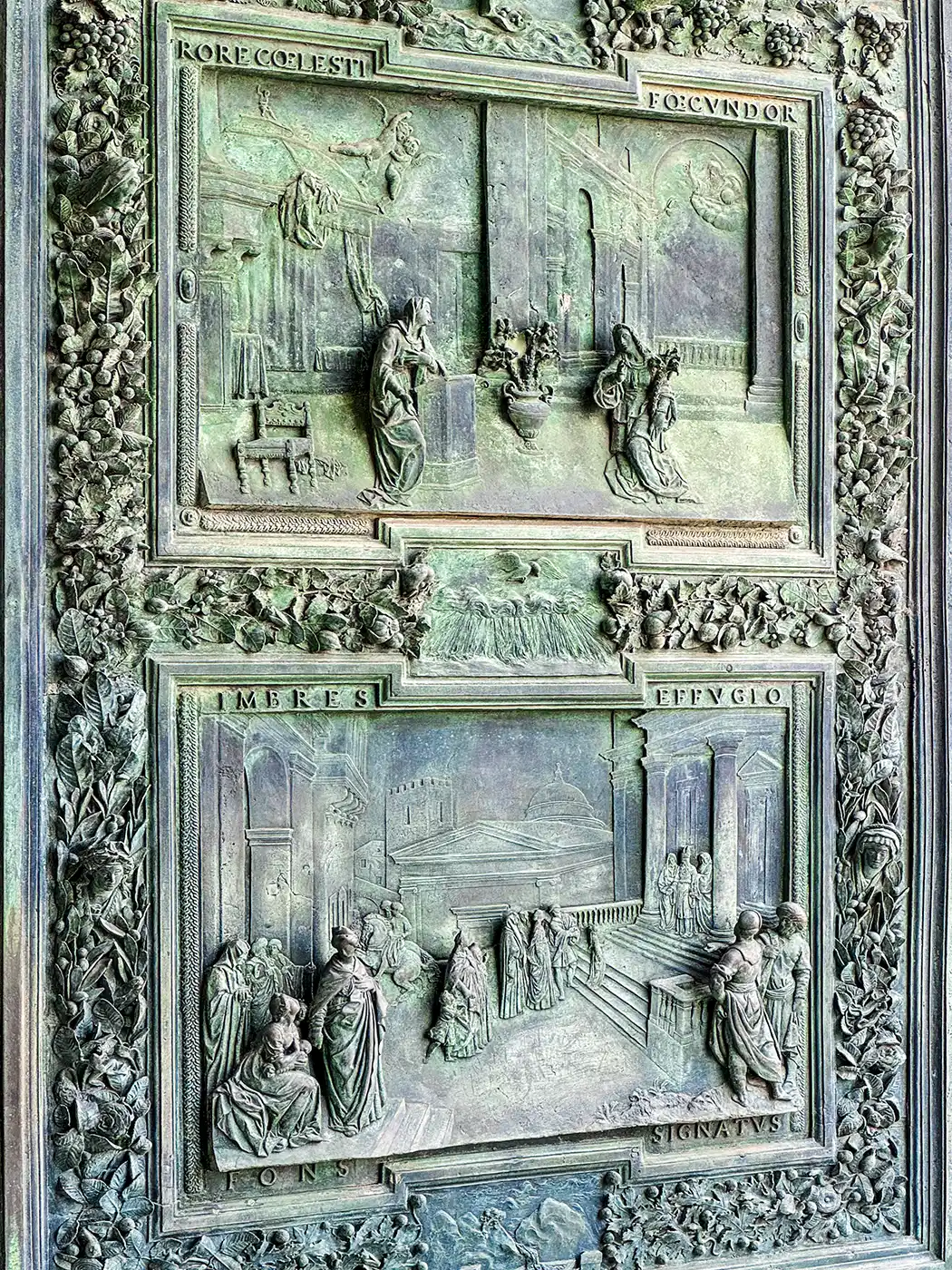 Türfüllungen aus Bronze mit detailreichen religiösen Darstellungen und lateinischen Inschriften, umrahmt von kunstvollen Blumenbordüren.
