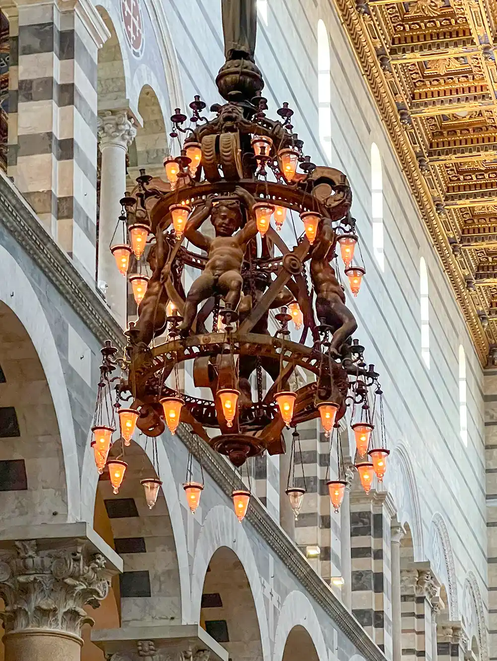 Ein kunstvoll verzierter Kronleuchter mit Figuren und leuchtenden Glühbirnen hängt in einem prächtigen Saal mit gestreiften Steinbögen und einer goldenen Decke.