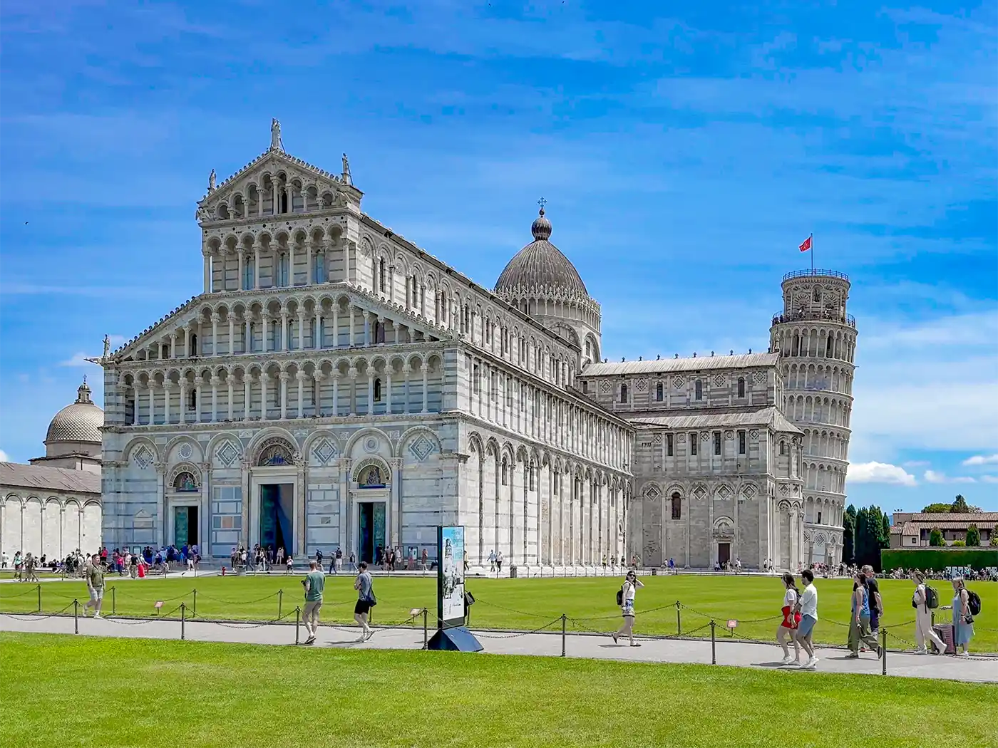 Der Schiefe Turm von Pisa und der Dom von Pisa an einem sonnigen Tag, mit Menschen, die auf dem Rasen spazieren gehen.