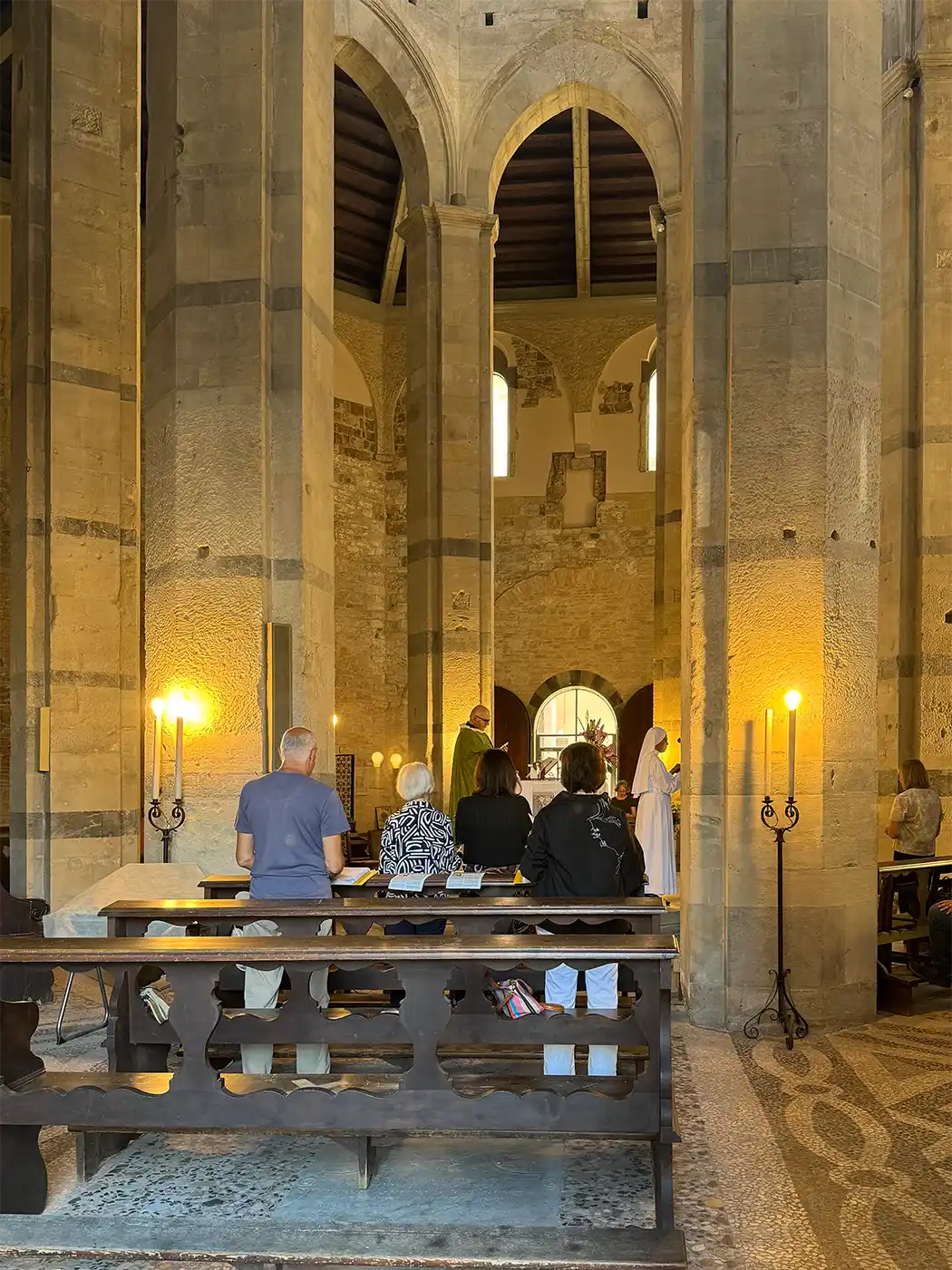 Pisa-Geheimtipp-Chiesa-del-Santo-Sepolcro-Gottesdienst In einer von Kerzen beleuchteten Steinkirche mit Gewölben und hohen Säulen sitzen und stehen Menschen und blicken auf einen Priester.