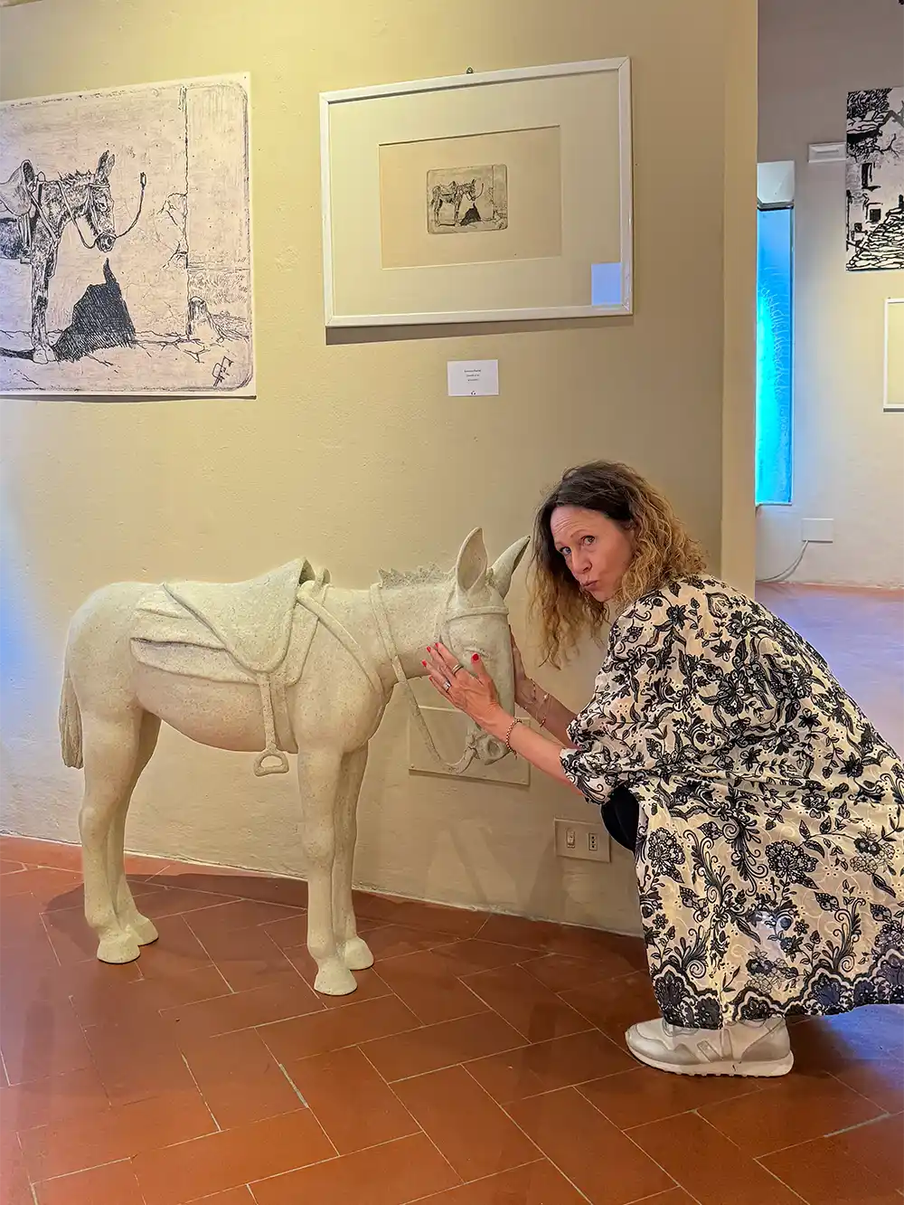Eine Frau hockt in einer Kunstgalerie neben einer kleinen weißen Eselstatue und streicht ihr sanft über den Kopf.