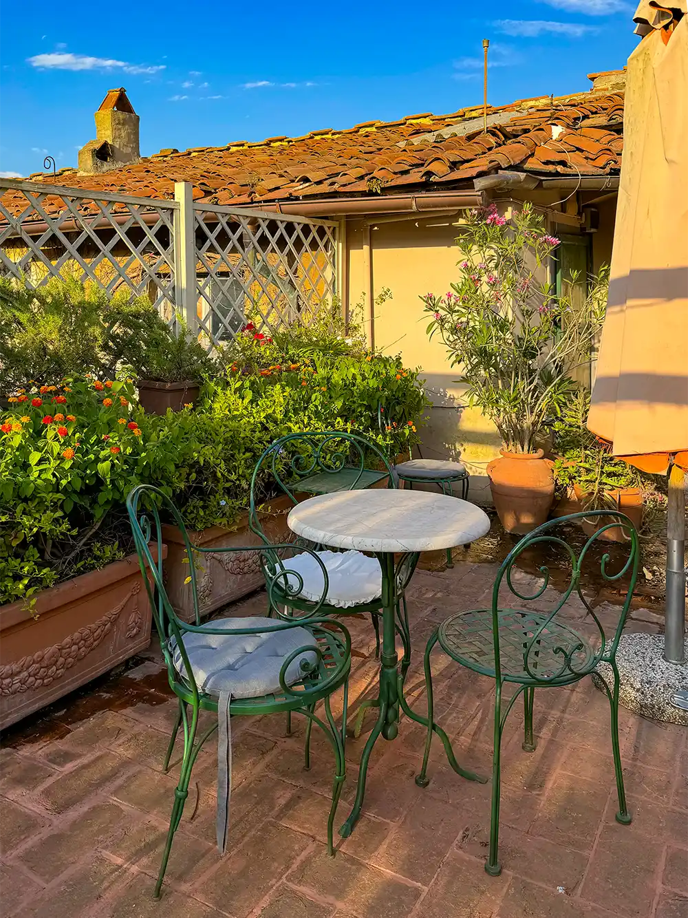 Pisa-Geheimtipp-Hotel-Royal-Victoria-Dachterrasse Ein kleiner runder Tisch mit drei Stühlen auf einer sonnigen Terrasse, umgeben von Topfpflanzen und Blumen.
