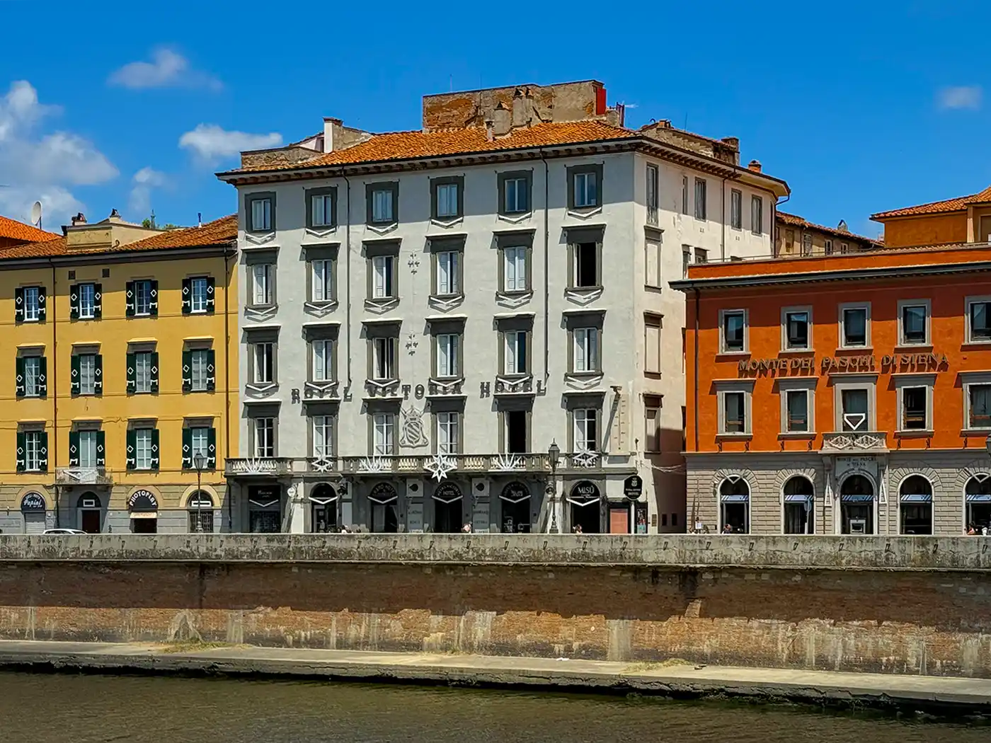 Pisa-Geheimtipp-Hotel-Royal-Victoria Unter einem strahlend blauen Himmel mit vereinzelten Wolken säumen farbenfrohe historische Gebäude eine Straße am Flussufer.