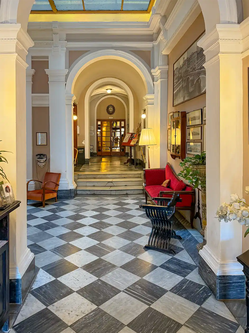 Pisa-Geheimtipp-Hotel-Victoria Elegante Hotellobby mit kariertem Boden, rotem Sofa, Bögen und warmer Beleuchtung, die in einen Flur führt.