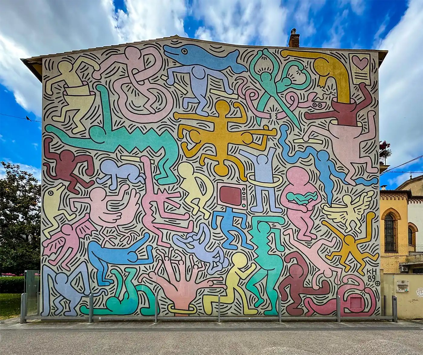 Pisa-Geheimtipp-Keith-Haring Farbenfrohes Wandgemälde mit abstrakten, konturierten Menschen-, Tier- und Herzformen, das eine gesamte Gebäudewand bedeckt.