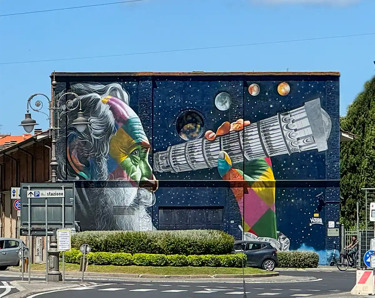 Pisa-Geheimtipps-Galileo-Galilei Farbenfrohes Wandbild, das einen bärtigen Mann zeigt, der durch ein Teleskop auf Planeten blickt, vor dem Hintergrund einer dunkelblauen, sternenübersäten Wand.