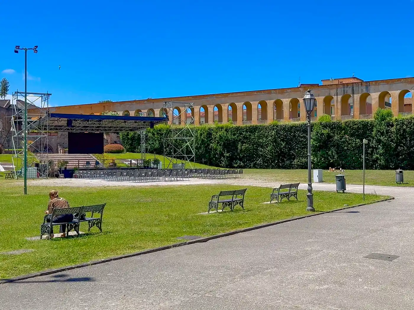 Eine Person sitzt auf einer Bank in einem sonnigen Park, im Hintergrund sind ein Aquädukt und eine Freiluftbühne zu sehen.