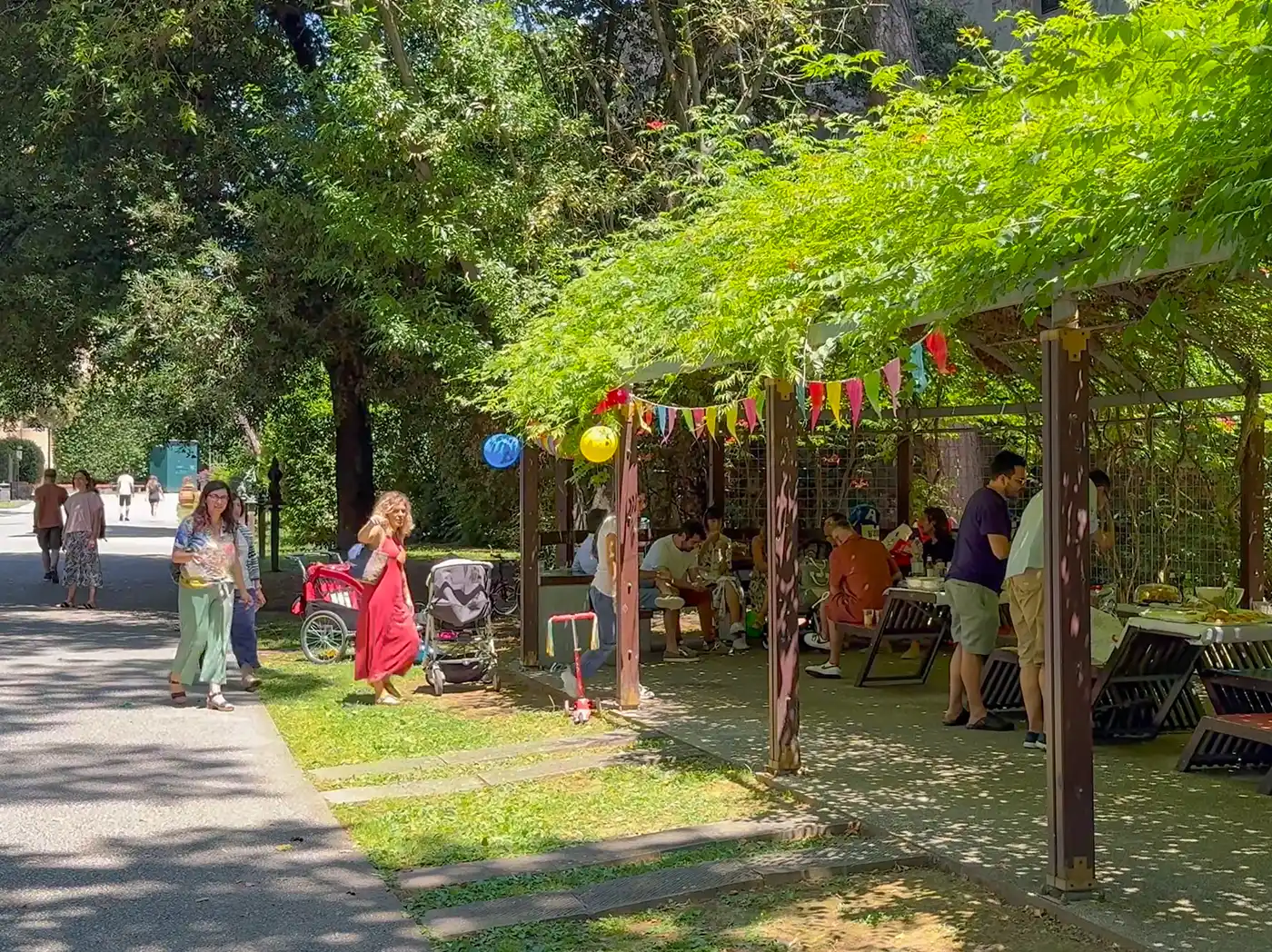 Unter einer geschmückten Pergola versammeln sich Menschen zu einer Feier; andere spazieren auf einem schattigen Parkweg in der Nähe.