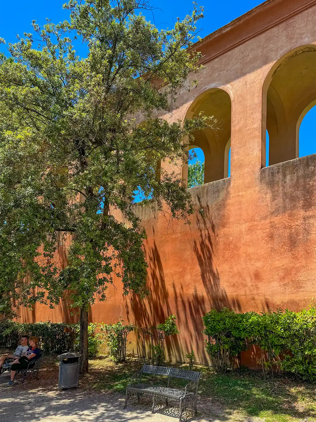Eine hohe orangefarbene Mauer mit Bögen, ein belaubter Baum, Bänke und zwei Personen, die links im Schatten sitzen.