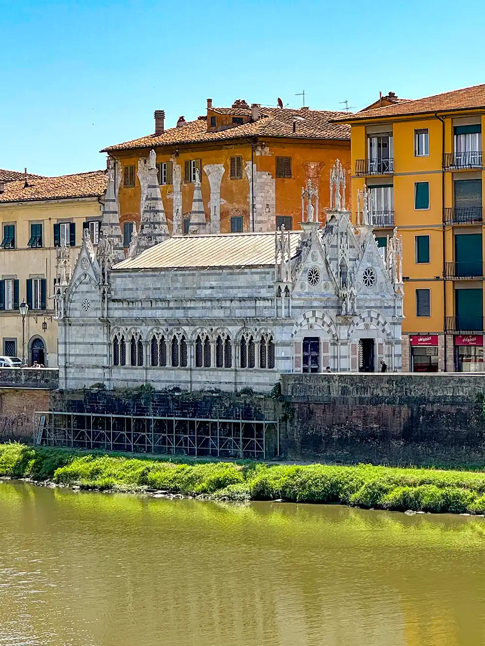 Pisa-Kirche Am Ufer eines Flusses steht eine kleine weiße gotische Kirche, im Hintergrund sind gelbe und ziegelrote Gebäude zu sehen.