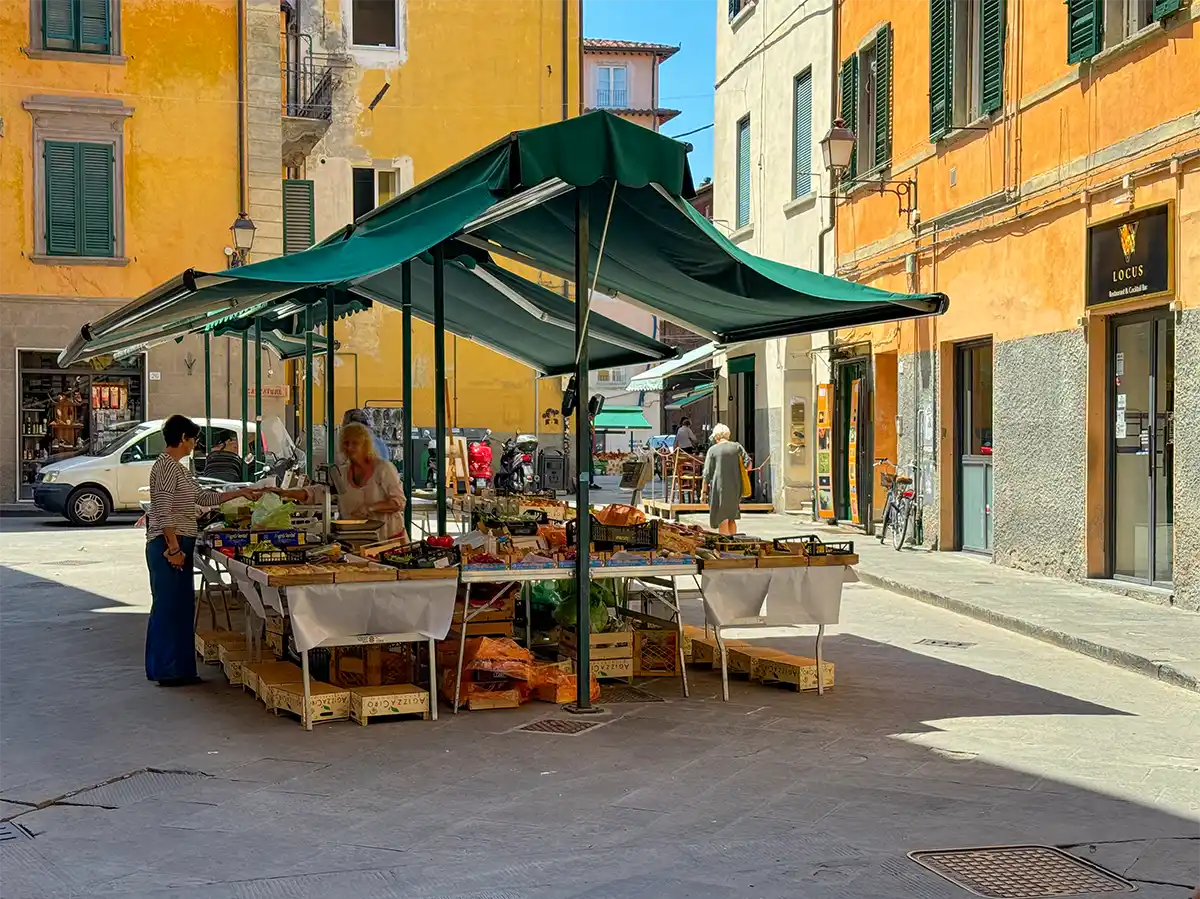 Pisa-Markt Ein Marktstand im Freien mit Obst und Gemüse, zwei Frauen beim Einkaufen, umgeben von bunten Gebäuden.