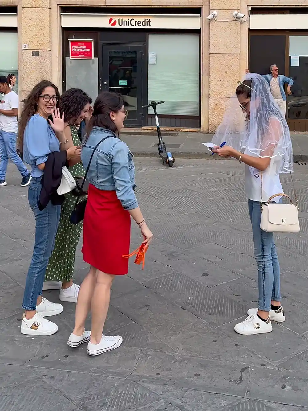Vier Frauen stehen auf einer Straße; eine trägt einen Schleier und schreibt, während die anderen lächeln und eine in die Kamera winkt.