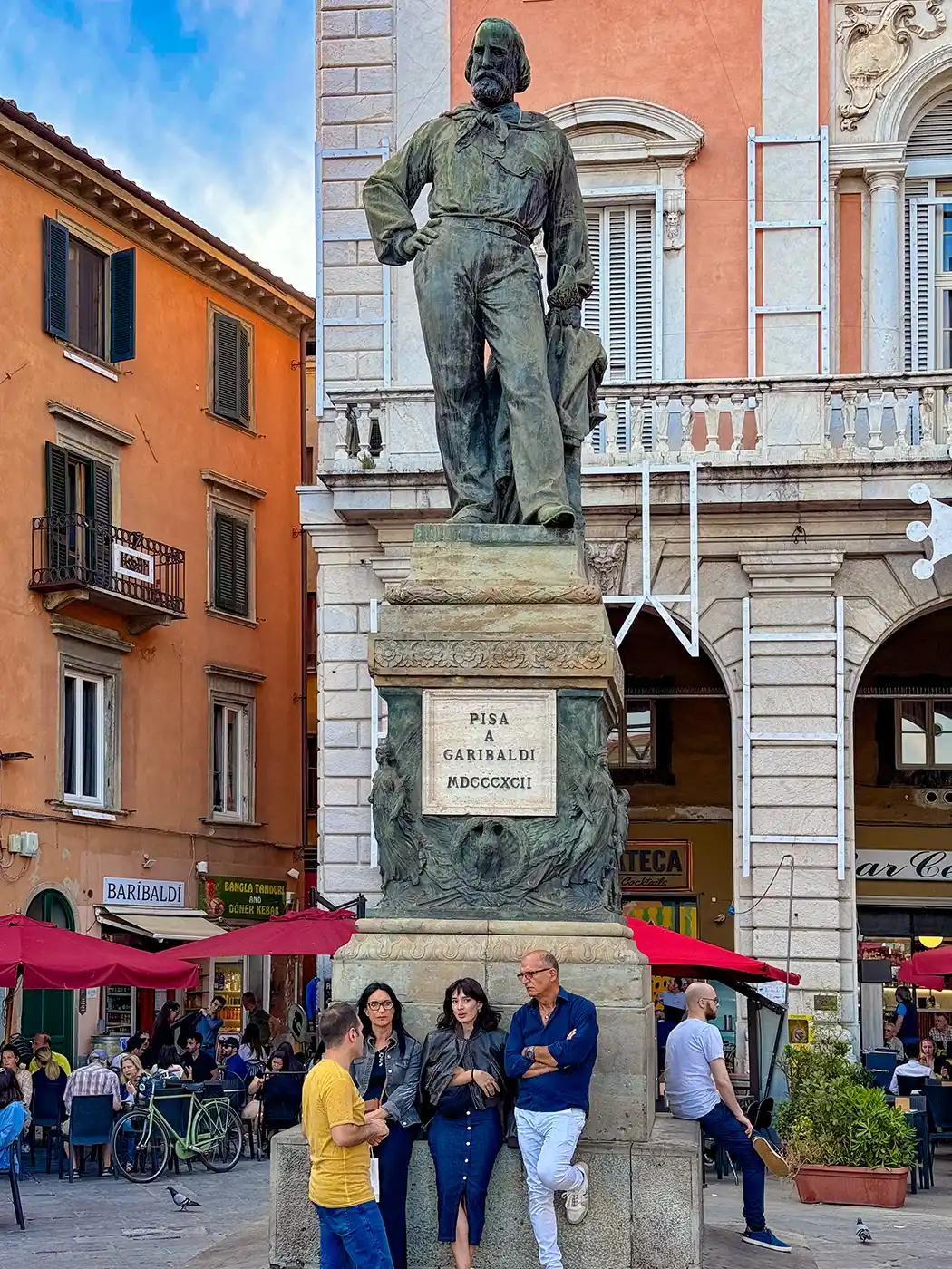 Vier Menschen stehen am Sockel einer großen Statue im Freien auf einem belebten europäischen Platz mit Cafés.