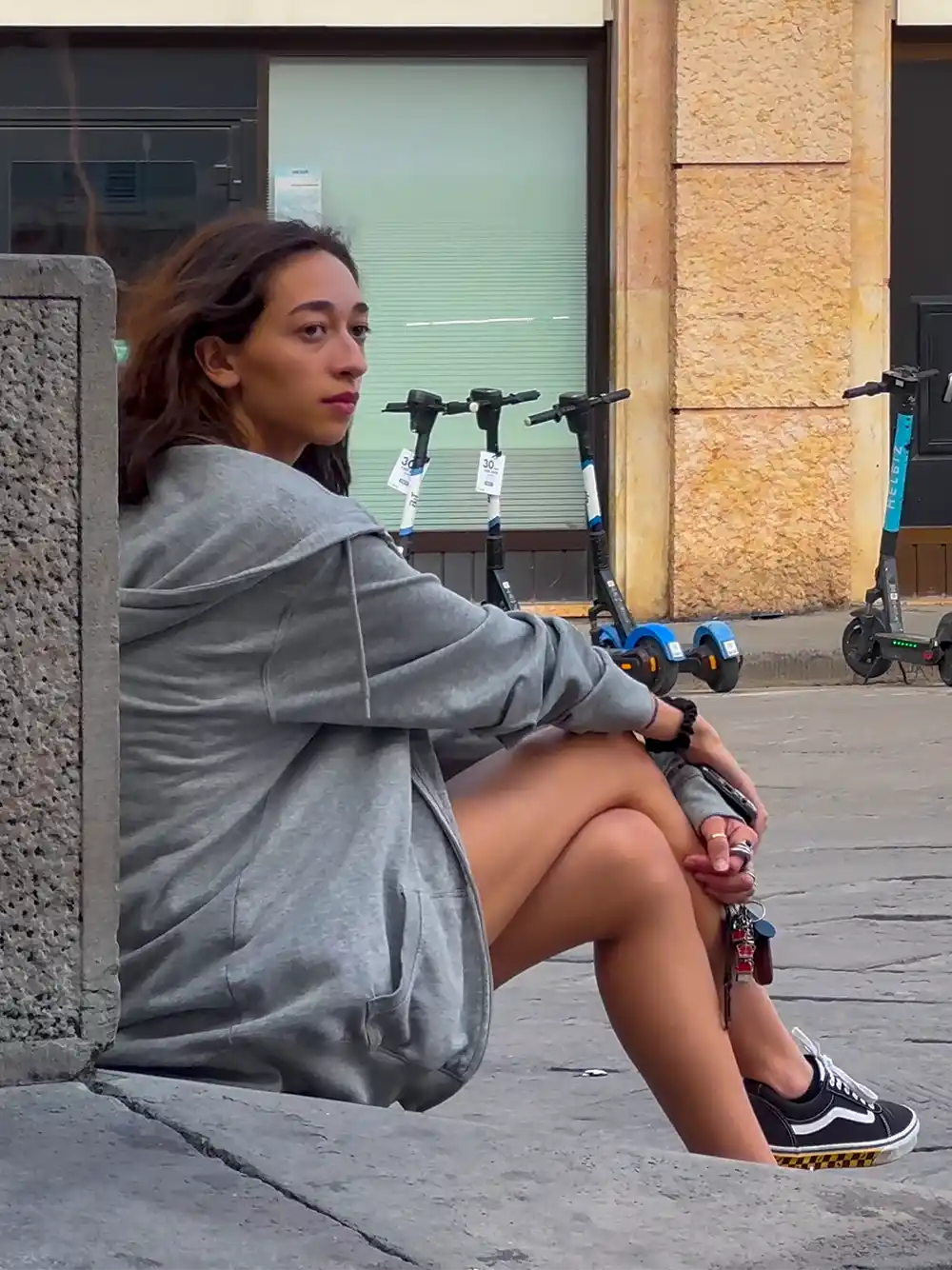 Eine Frau in einem grauen Kapuzenpulli sitzt auf dem Bürgersteig und hält einen Roller in der Hand; im Hintergrund stehen E-Roller.