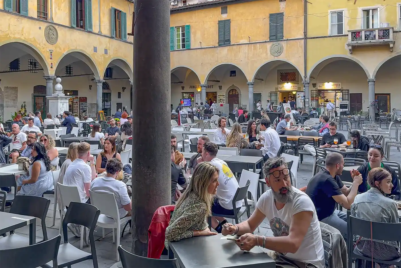 Menschen, die an Tischen im Freien in einem belebten italienischen Innenhof sitzen, im Hintergrund gelbe Gebäude und Bögen.