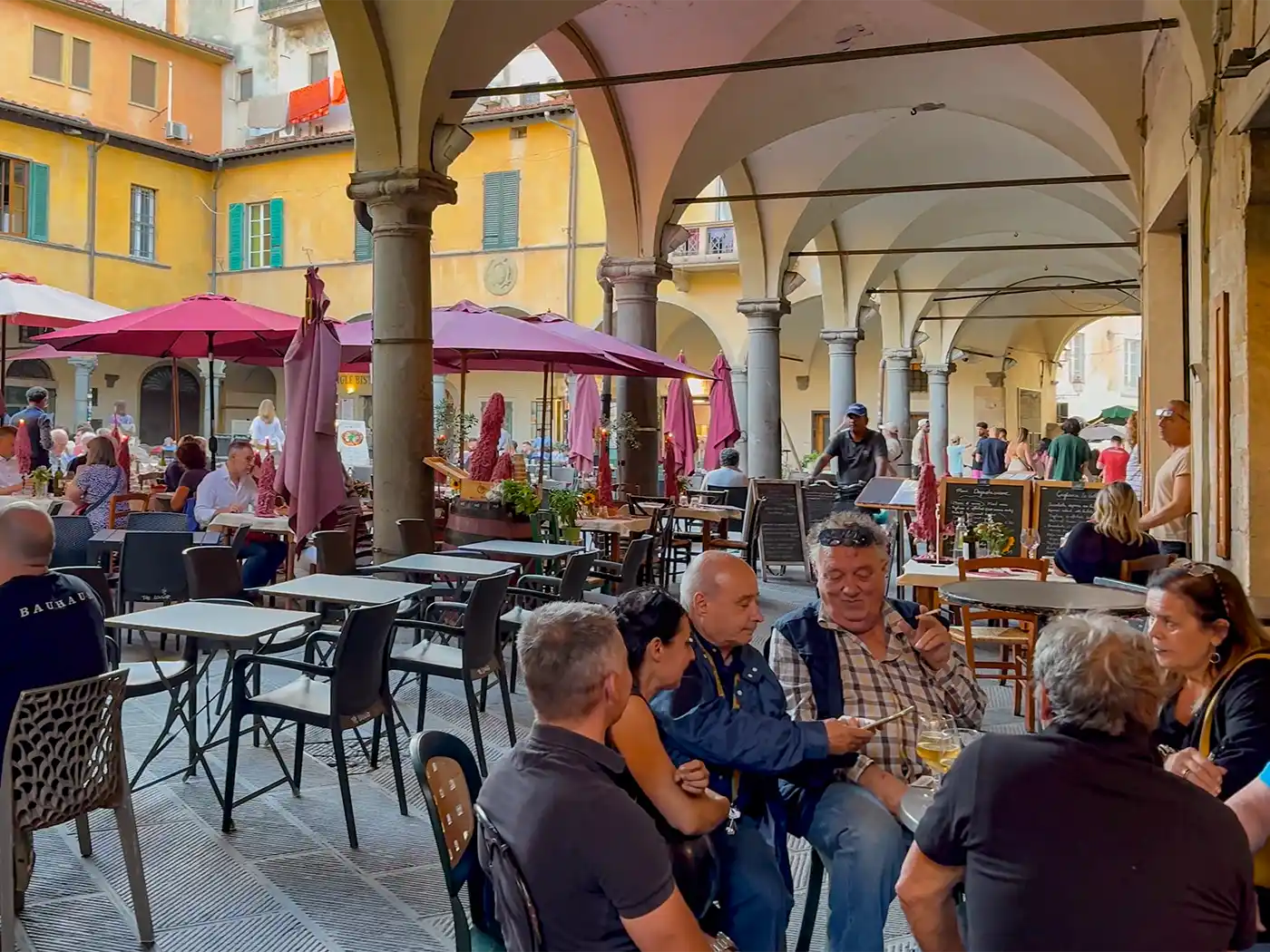 Pisa-Piazza-Vettovaglie Auf einem belebten Platz mit gelben Gebäuden und roten Sonnenschirmen sitzen Menschen an Tischen im Freien unter Arkaden.