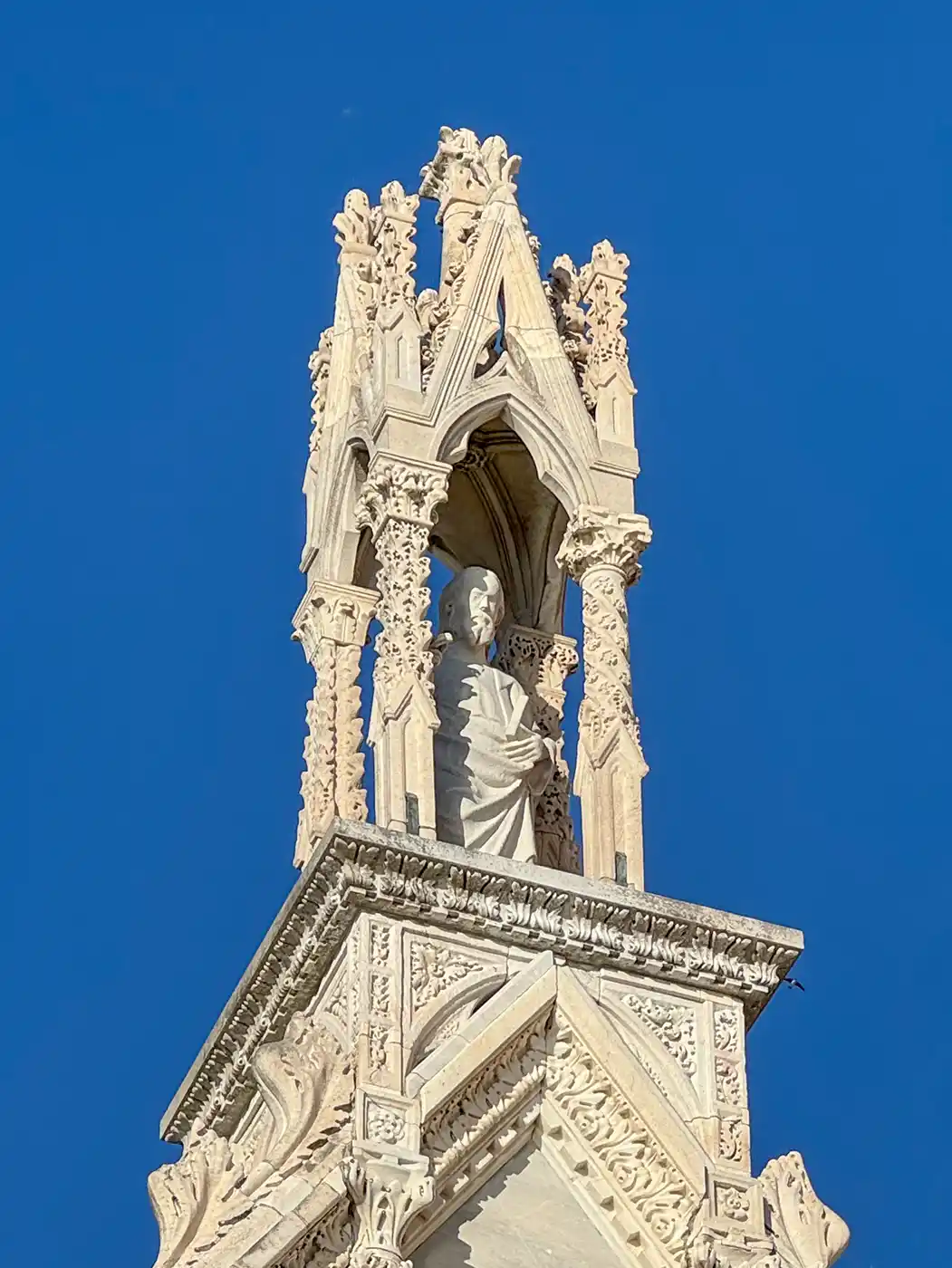 Eine Statue aus weißem Marmor unter einem kunstvollen gotischen Baldachin auf dem Dach eines Gebäudes vor strahlend blauem Himmel.