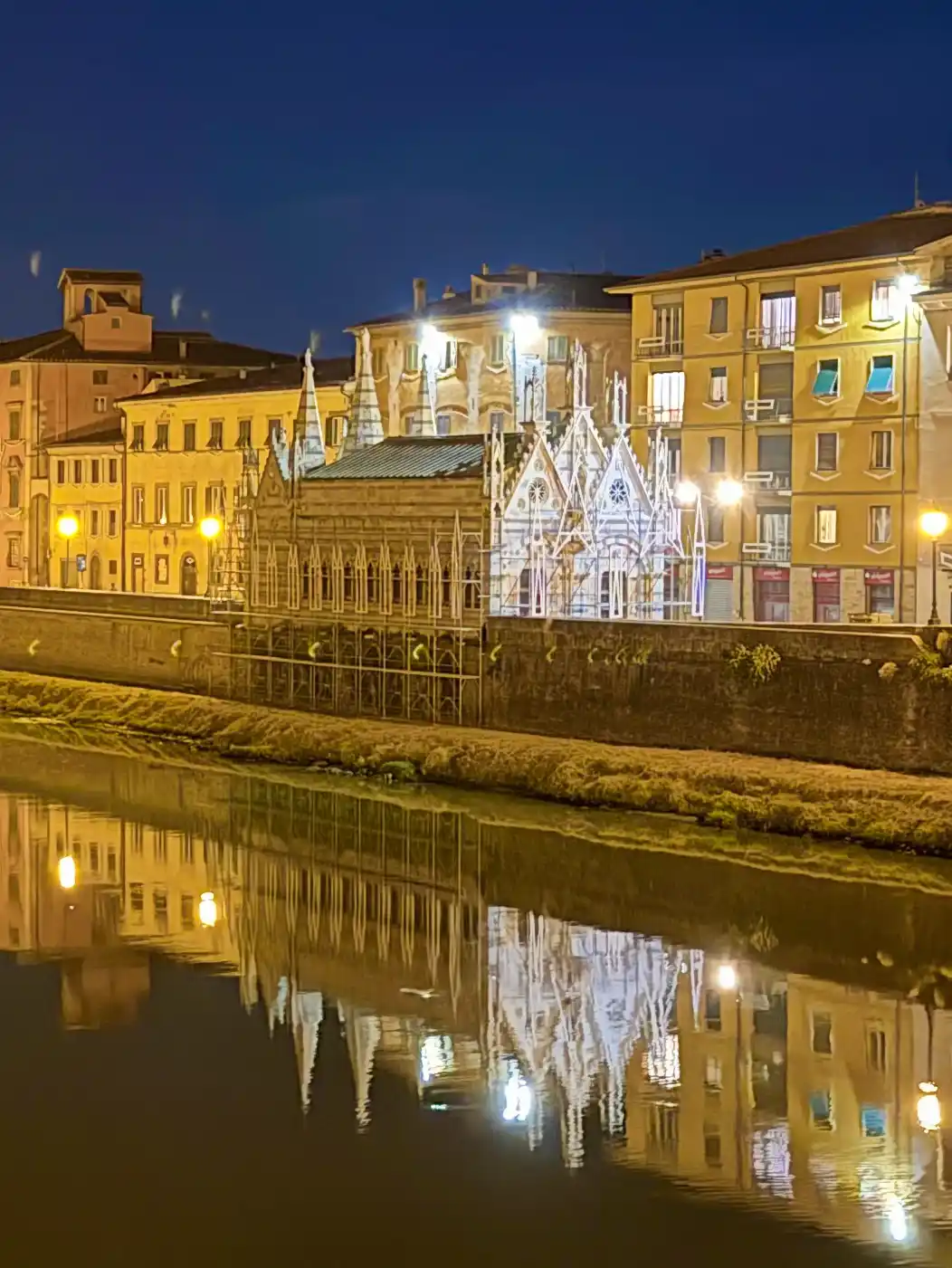 Eine Kirche im gotischen Stil und farbenfrohe Gebäude, die sich nachts in einem Fluss spiegeln und von warmem Licht beleuchtet werden.