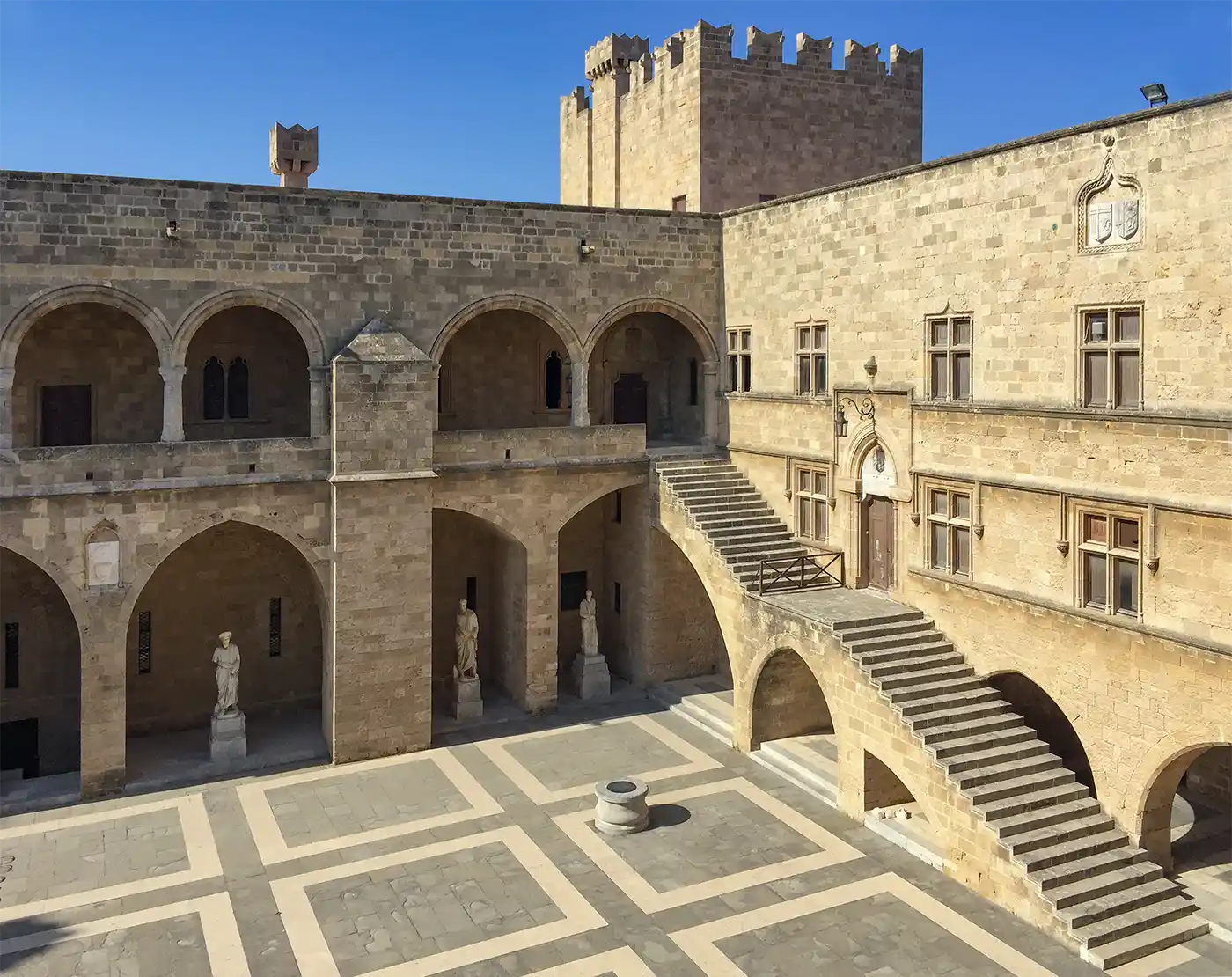 Ein steinerner Innenhof mit Bögen, Statuen, einer Treppe und einem Schlossturm unter strahlend blauem Himmel.