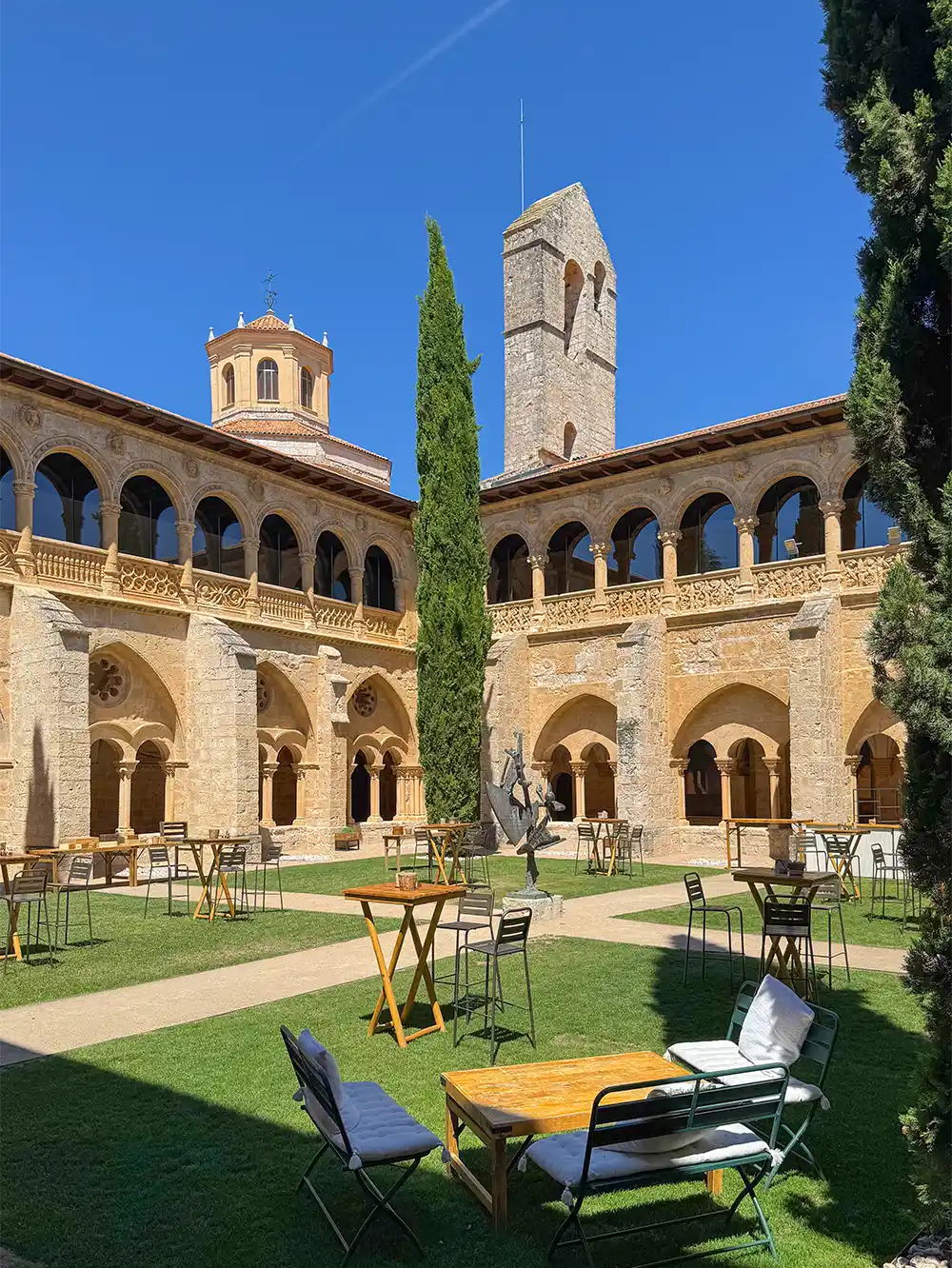 Valbuena-Castilla-Termal Ein mit Steinen gepflasterter Innenhof mit Bögen, Sitzgelegenheiten im Freien, grünem Rasen, hohen Zypressen und einem blauen Himmel darüber.