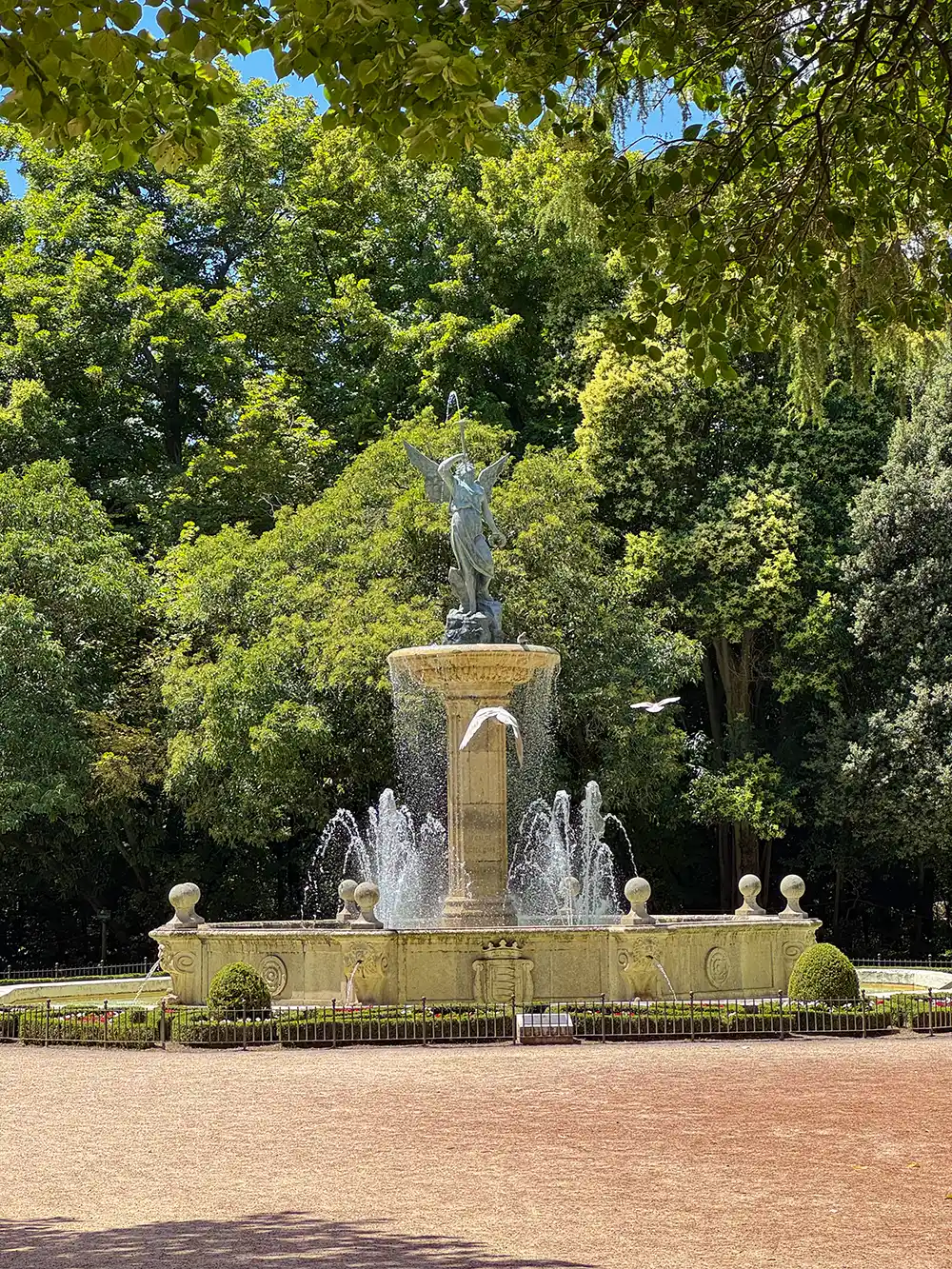 Valladolid-Campo-Grande-Brunnen Ein Springbrunnen mit einer geflügelten Statue und Wasserstrahlen, umgeben von Bäumen in einem sonnigen Park.