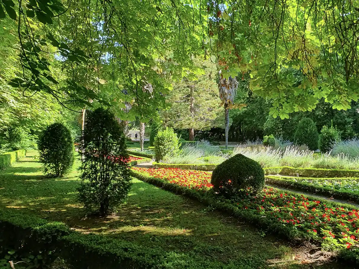 Valladolid-Campo-Grande-Garten Ein sonnendurchfluteter Garten mit gepflegten Sträuchern, bunten Blumenbeeten und üppig grünen Bäumen, die reichlich Schatten spenden.