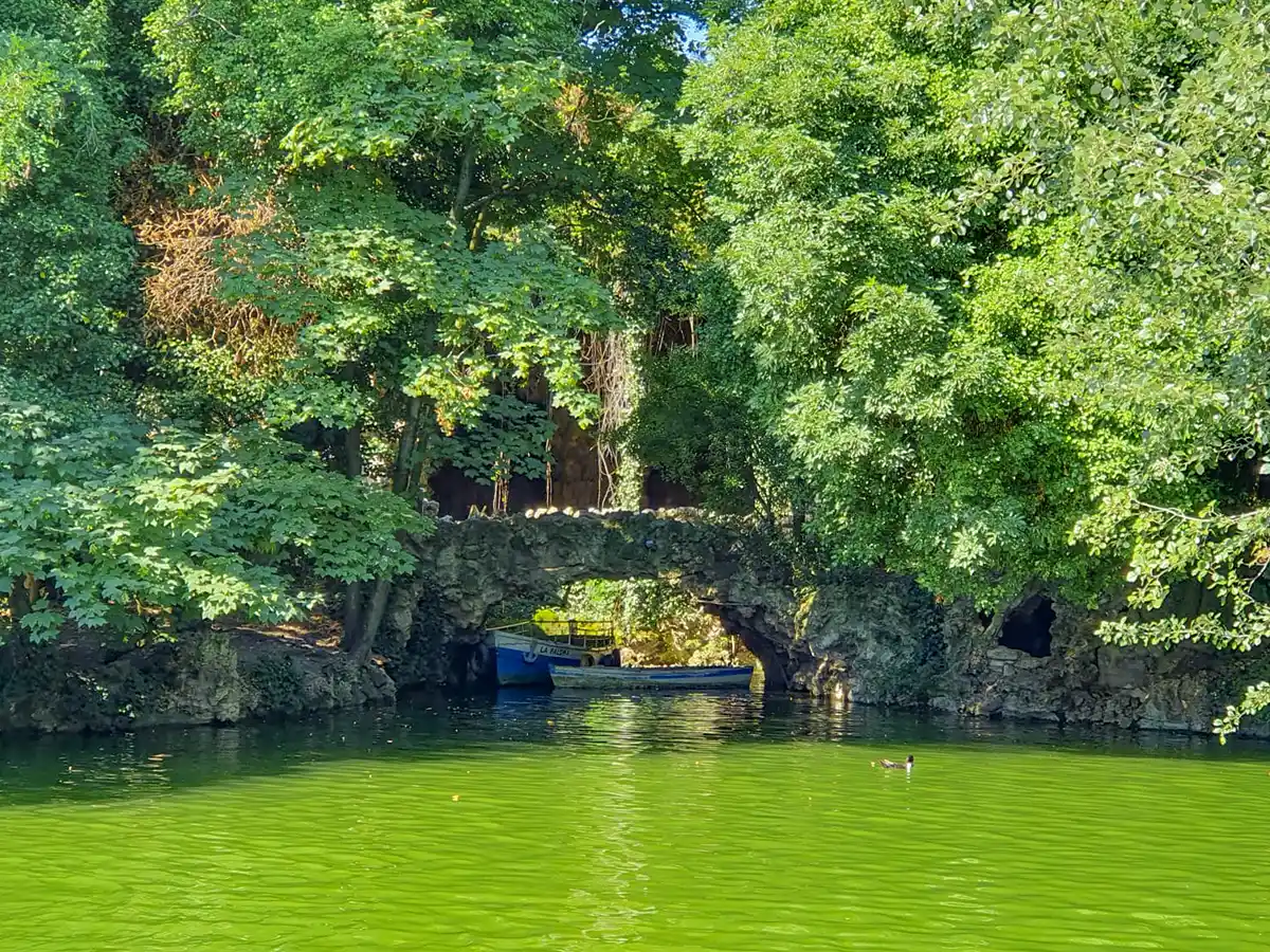 Valladolid-Campo-Grande-Teich Eine kleine Steinbrücke über einem grünen Teich, umgeben von üppigen Bäumen, im Hintergrund ist ein blaues Boot zu sehen.