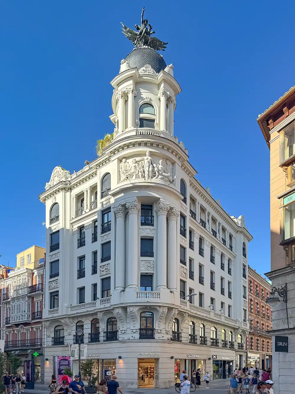 Valladolid-Casa-Fenix Weißes historisches Gebäude mit Säulen und einer Kuppel, gelegen an einer belebten Straße, an der Menschen vorbeigehen.