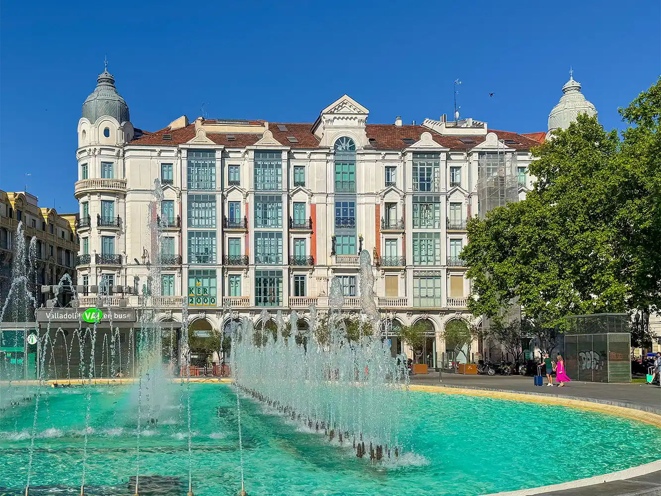 Valladolid-Casa-Mantilla Ein Springbrunnen vor einem prächtigen Gebäude mit Glasfenstern, Bäumen und einem strahlend blauen Himmel.
