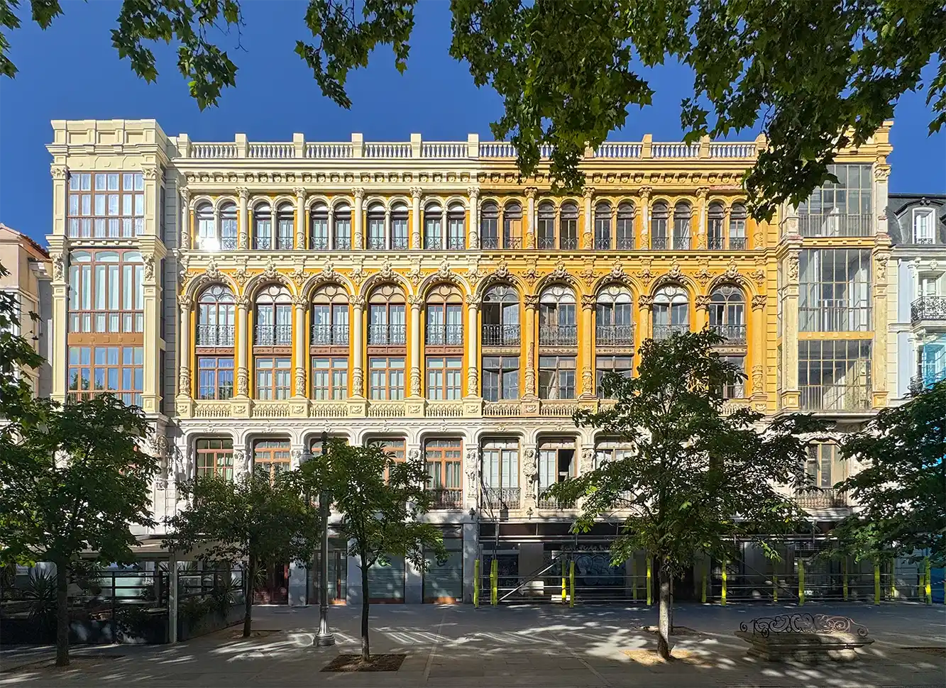 Valladolid-Casa-Resines Ein historisches, prunkvolles gelbes Gebäude mit Bogenfenstern, das an einem sonnigen Tag hinter Laubbäumen zu sehen ist.