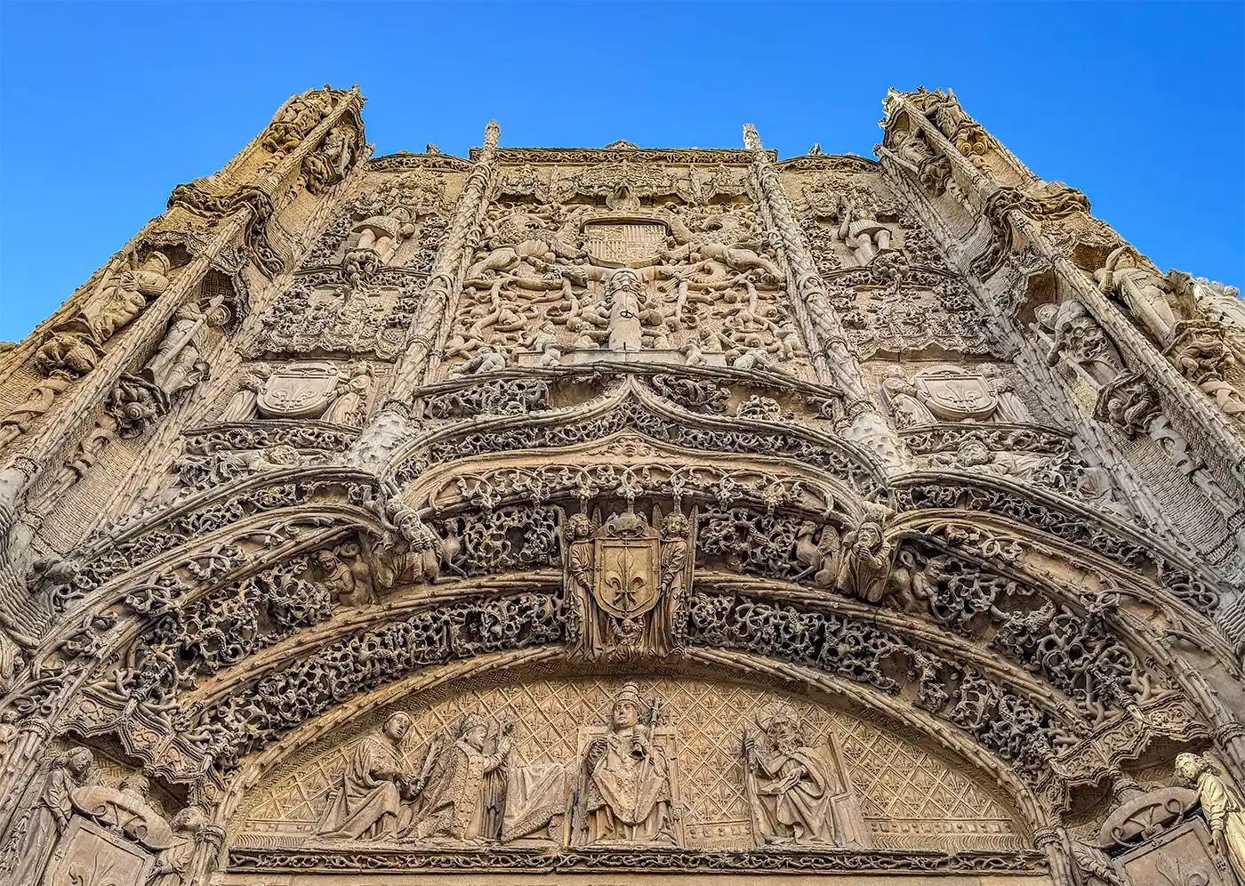 Valladolid-Colegio-San-Gregorio-Fassade Die kunstvoll verzierte Steinfassade eines historischen Gebäudes mit detailreichen Figuren, von unten vor blauem Himmel betrachtet.