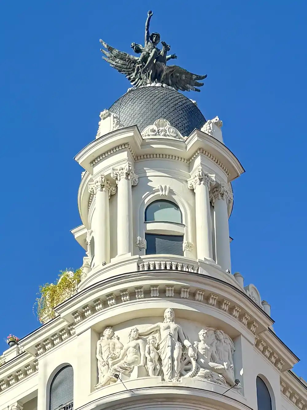 Valladolid-Fenix-Detail-2 Ein weißes, kuppelförmiges Gebäude mit detailreichen Skulpturen und einer geflügelten Statue auf der Spitze, vor dem Hintergrund eines strahlend blauen Himmels.