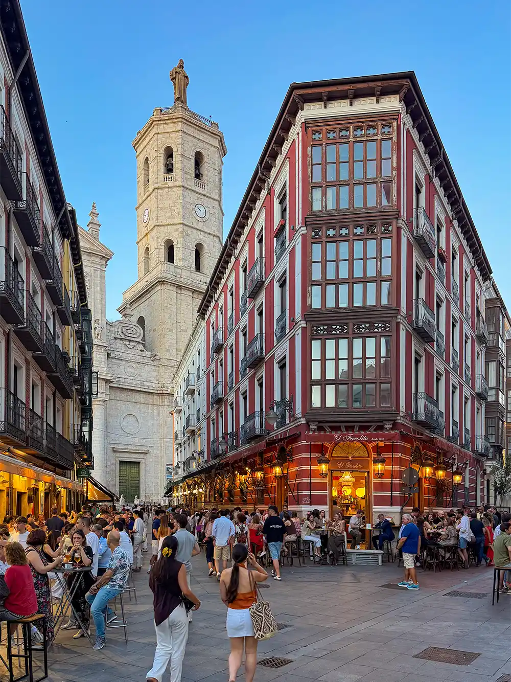Valladolid-Kathedrale-Calle-Gascajares Menschen essen an Tischen im Freien in der Nähe eines roten, historischen Gebäudes und eines hohen Kirchturms auf einer belebten Stadtstraße.