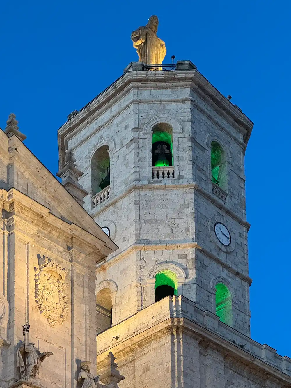 Valladolid-Kathedrale-Turm Ein Uhrturm aus Stein mit grün beleuchteten Fenstern und einer goldenen Statue auf der Spitze, vor dem Hintergrund eines strahlend blauen Himmels.
