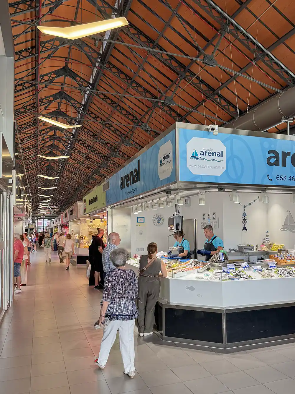 Valladolid-Mercado-de-Val-innen Hallenmarkt, auf dem Menschen an Verkaufsständen unter einem hohen, mit orangefarbenen Fliesen gedeckten Dach mit freiliegenden Balken einkaufen.