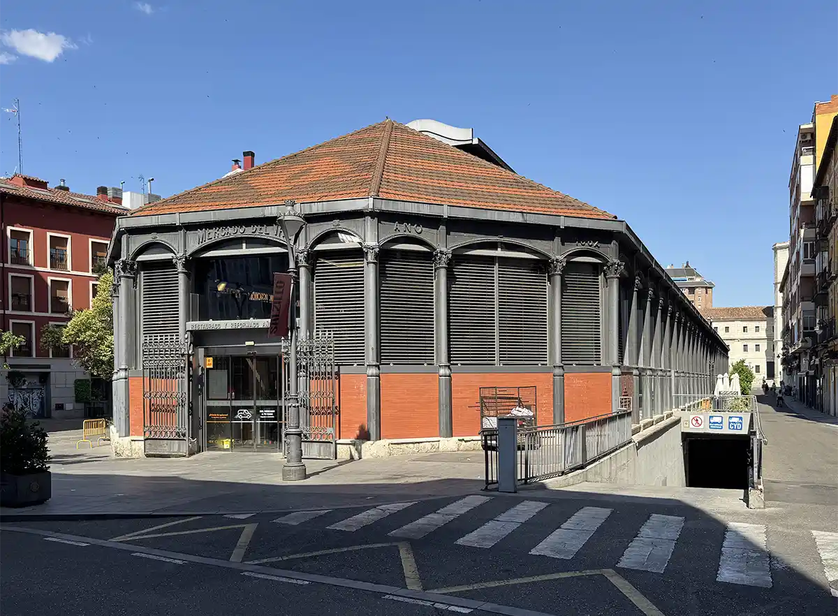 Valladolid-Mercado-de-Val Ein Marktgebäude aus Metall und Ziegeln mit einem schrägen roten Dach an einer Straßenecke unter strahlend blauem Himmel.