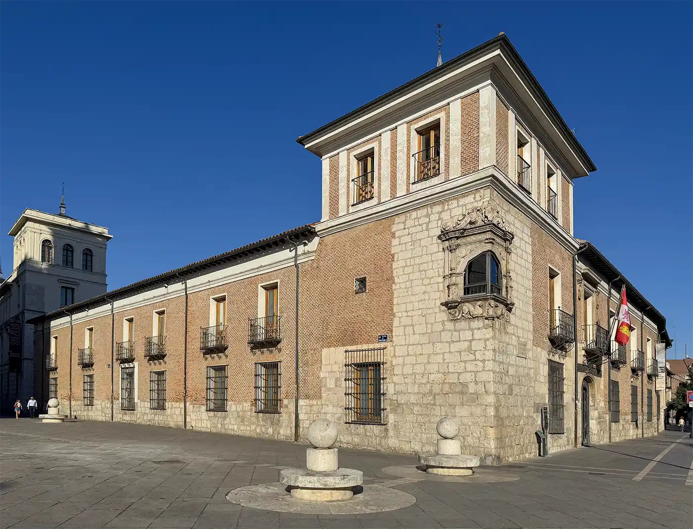 Valladolid-Palacio-Pimentel Historisches Backstein- und Steingebäude mit einem Turm unter strahlend blauem Himmel, spanische Flaggen am Eingang.
