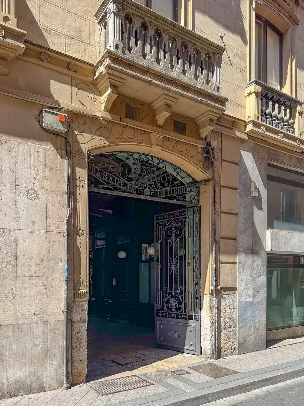 Valladolid-Pasaje-Gutierrez-Eingang Ein kunstvoll geschmiedetes Eisentor steht offen in einem alten Steinhaus, über dessen Eingang sich ein kleiner Balkon befindet.