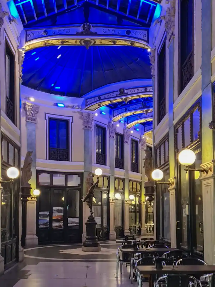 Valladolid-Pasaje-Gutierrez-abends Eine überdachte Arkade mit blau beleuchteter Glasdecke, verzierten Säulen, Statuen und leeren Cafétischen, die entlang des Gehwegs aufgereiht sind.