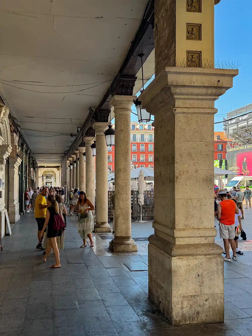 Valladolid-Plaza-Mayor-Arkaden Menschen spazieren unter einer überdachten Steinarkade mit Säulen entlang, die an einen sonnigen Platz in einem Stadtviertel grenzt.