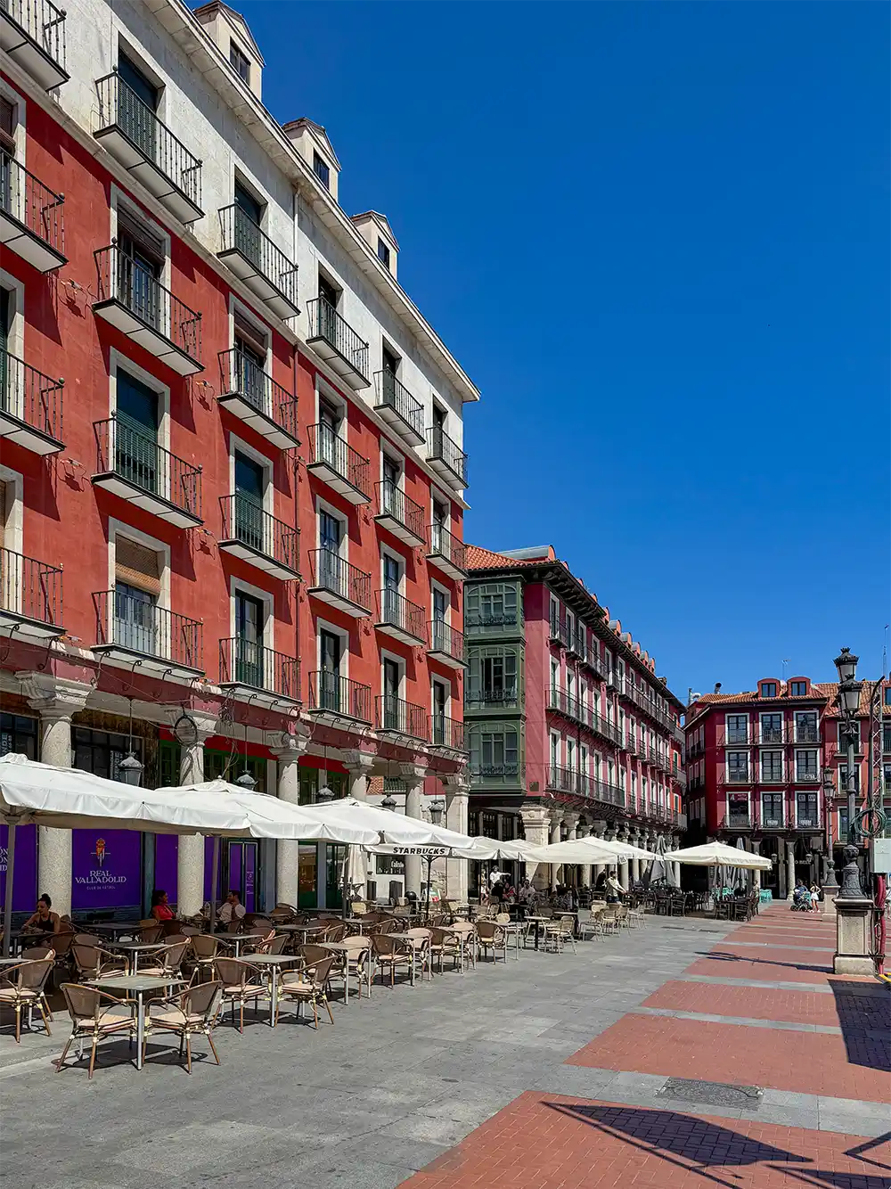 Valladolid-Plaza-Mayor-Fassaden Unter einem strahlend blauen Himmel säumen Café-Tische mit Sonnenschirmen einen Platz neben roten Gebäuden.